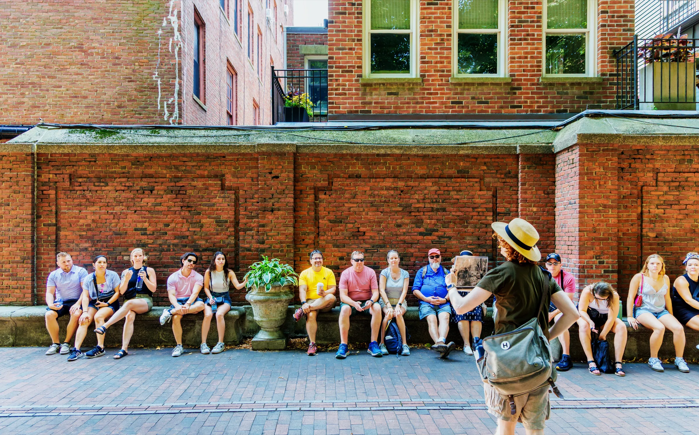 A guide in a straw hat speaking to a tour group seated along a brick wall