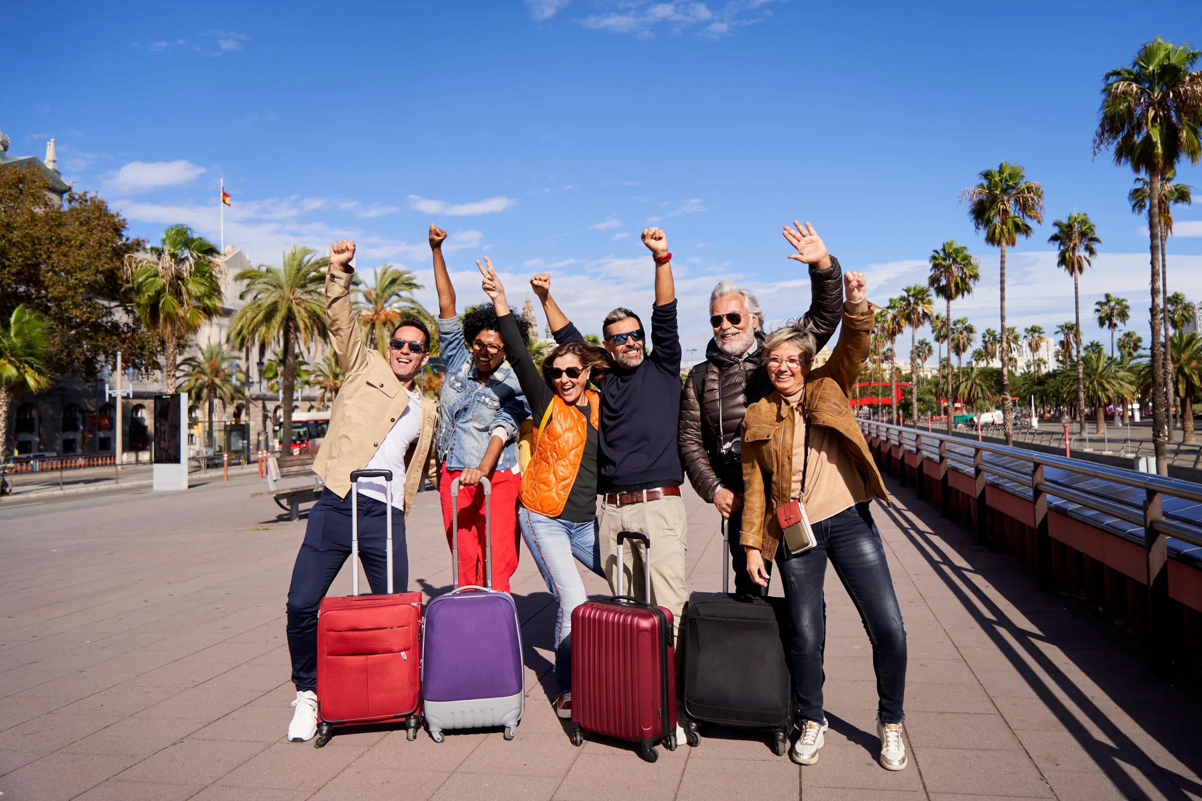 A group of adult travelers with their suitcases, cheering on a palm-lined promenade