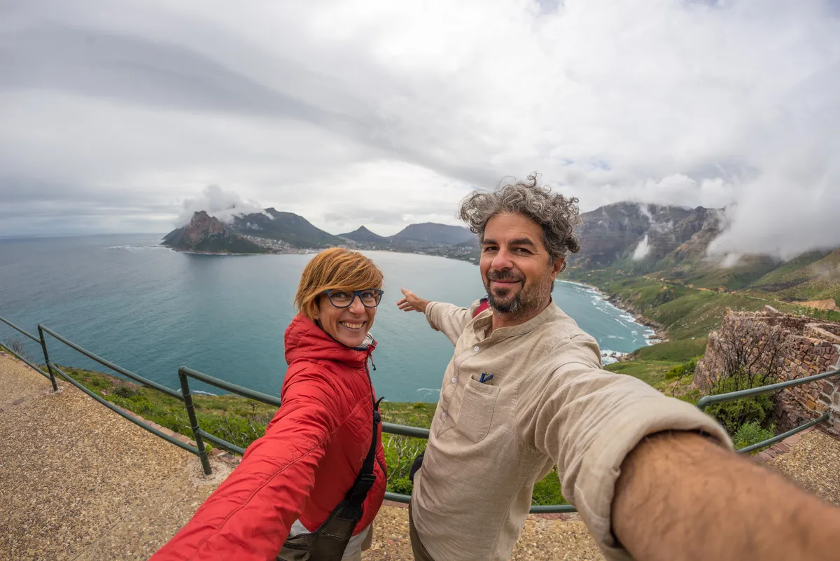 Two travelers taking a selfie above a bay on the Cape Peninsula, mountains in cloud behind