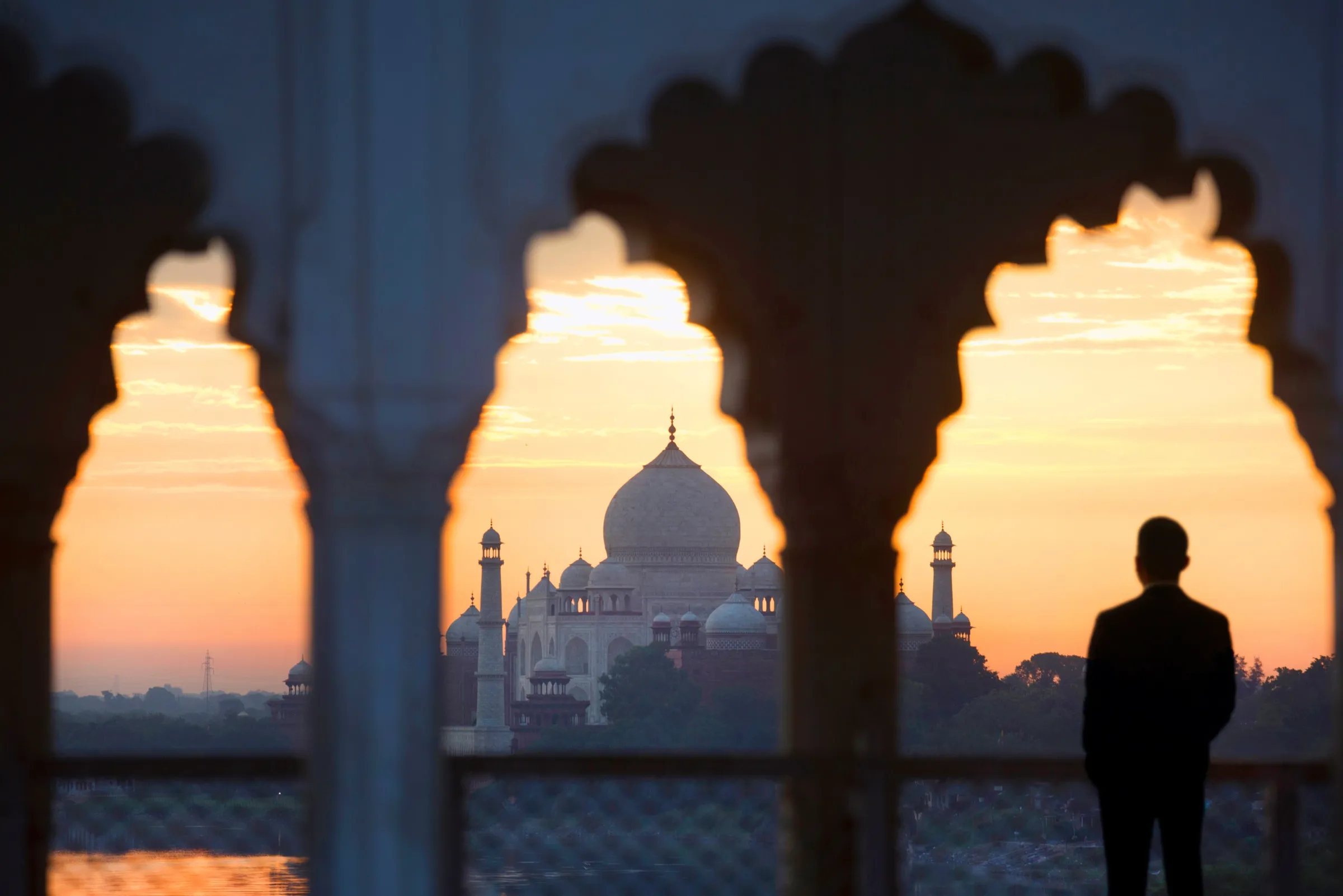The Taj Mahal at sunrise, framed by carved arches, with a traveler looking out