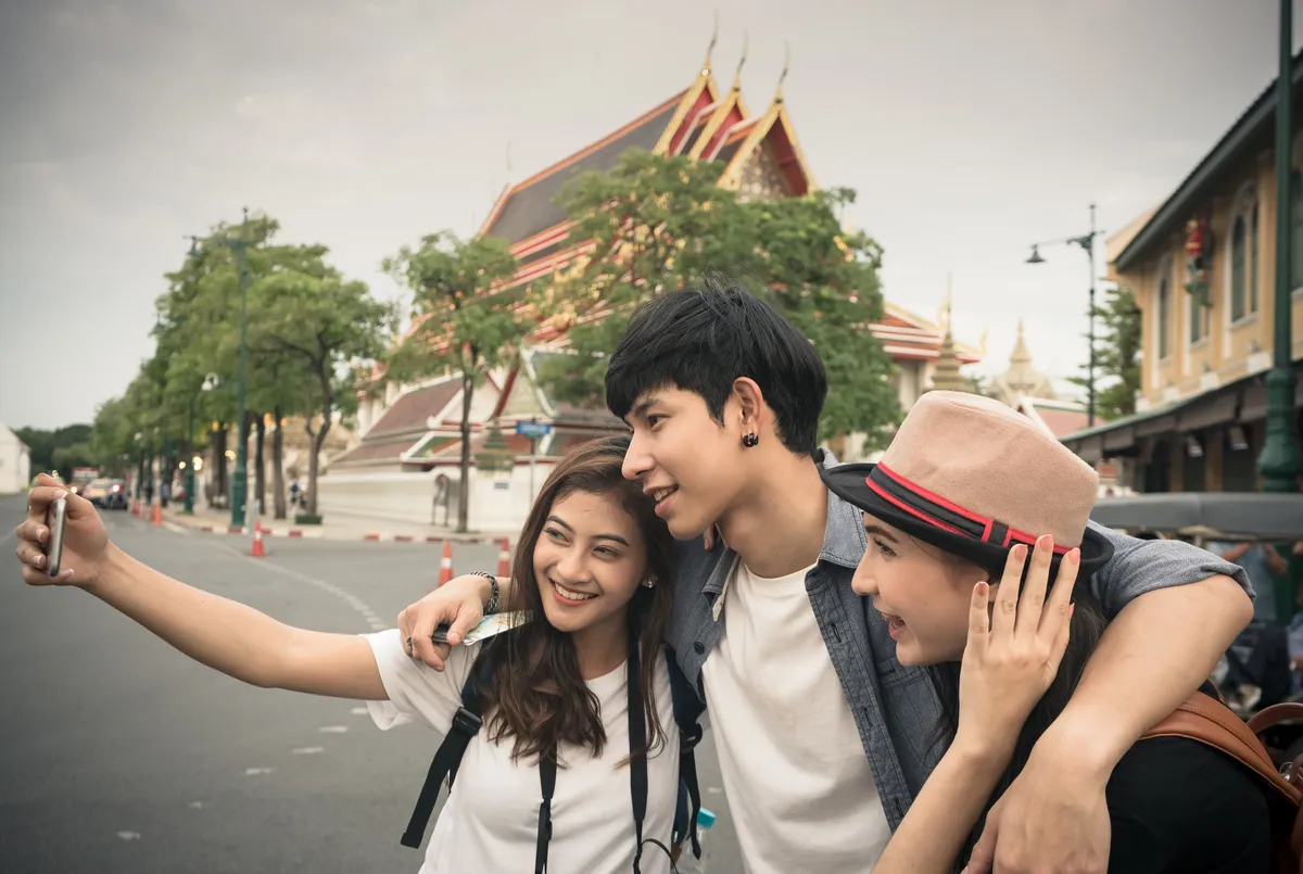 Three young travelers taking a selfie on a street beside a Bangkok temple with tiered red and gold roofs