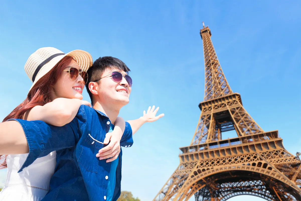 A smiling couple in sunglasses pointing up at the Eiffel Tower against a blue sky