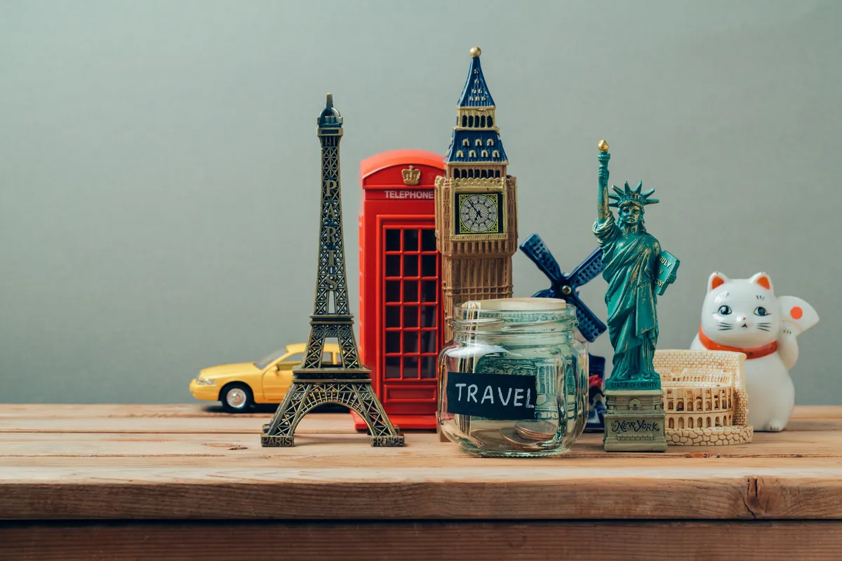 Souvenir landmarks on a wooden table, the Eiffel Tower, Big Ben, a red phone box and the Statue of Liberty, beside a jar of coins labeled Travel